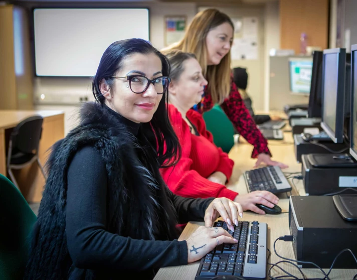 Student in a black vest working at a computer while a lecturer provides guidance and feedback in a classroom setting. Student in a black vest working at a computer while a lecturer provides guidance and feedback in a classroom setting.